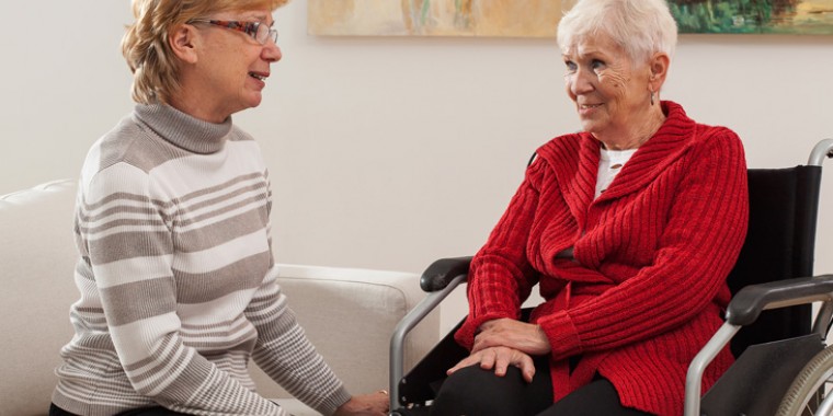 Two elderly women conversing in a room, one seated on a sofa and the other in a wheelchair. Both appear engaged and are next to a coffee table with tea cups on it.
