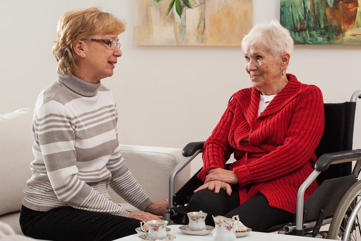 Two elderly women conversing in a room, one seated on a sofa and the other in a wheelchair. Both appear engaged and are next to a coffee table with tea cups on it.