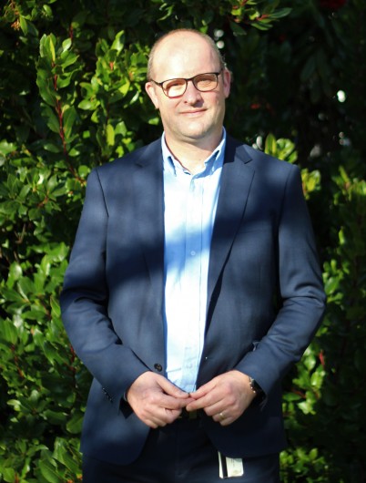 Man wearing glasses facing camera and smiling. He is standing outside in front of a shrub. 